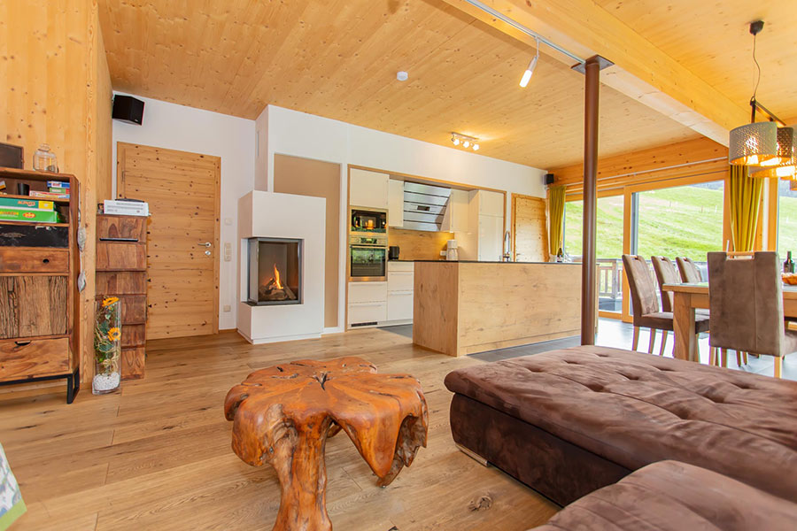 Offener Wohnraum mit Holzboden, braunem Sofa, rustikalem Baumstamm-Couchtisch, moderner Küche, Kamin und großem Fenster mit Blick auf eine grüne Wiese