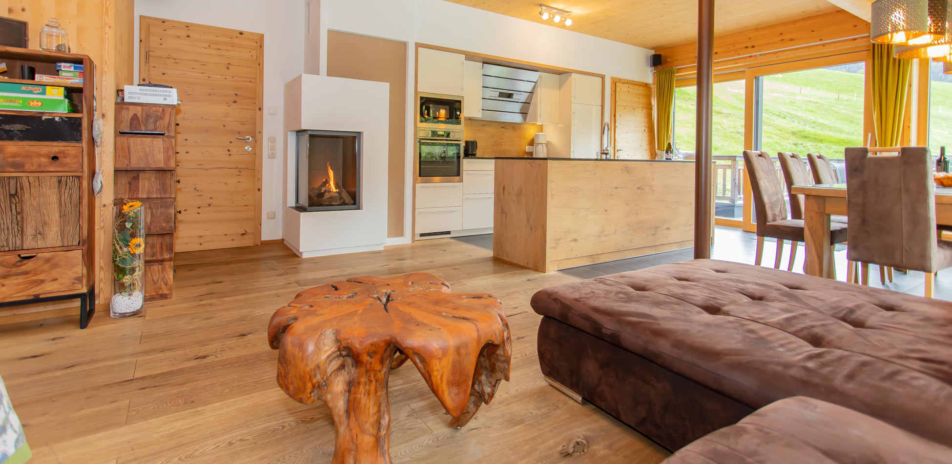 Innenraum eines Chalets in Saalbach‑Hinterglemm: Wohnbereich mit gemütlichem Sofa, modernem Kamin, offener Küche mit Kochinsel, Holzboden und großen Panoramafenstern mit Blick auf die grüne Berglandschaft