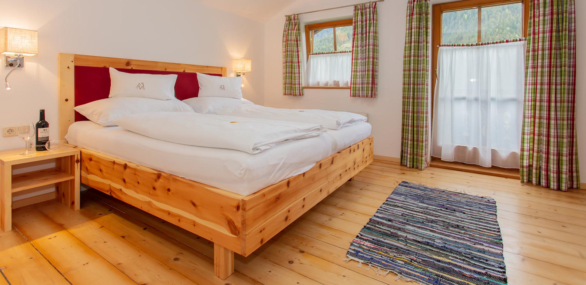 Helles Schlafzimmer in einem Chalet in Saalbach‑Hinterglemm mit einem gemütlichen Doppelbett auf Holzrahmen, Holzfußboden, karierten Vorhängen und Fenster mit Blick auf die Berglandschaft.