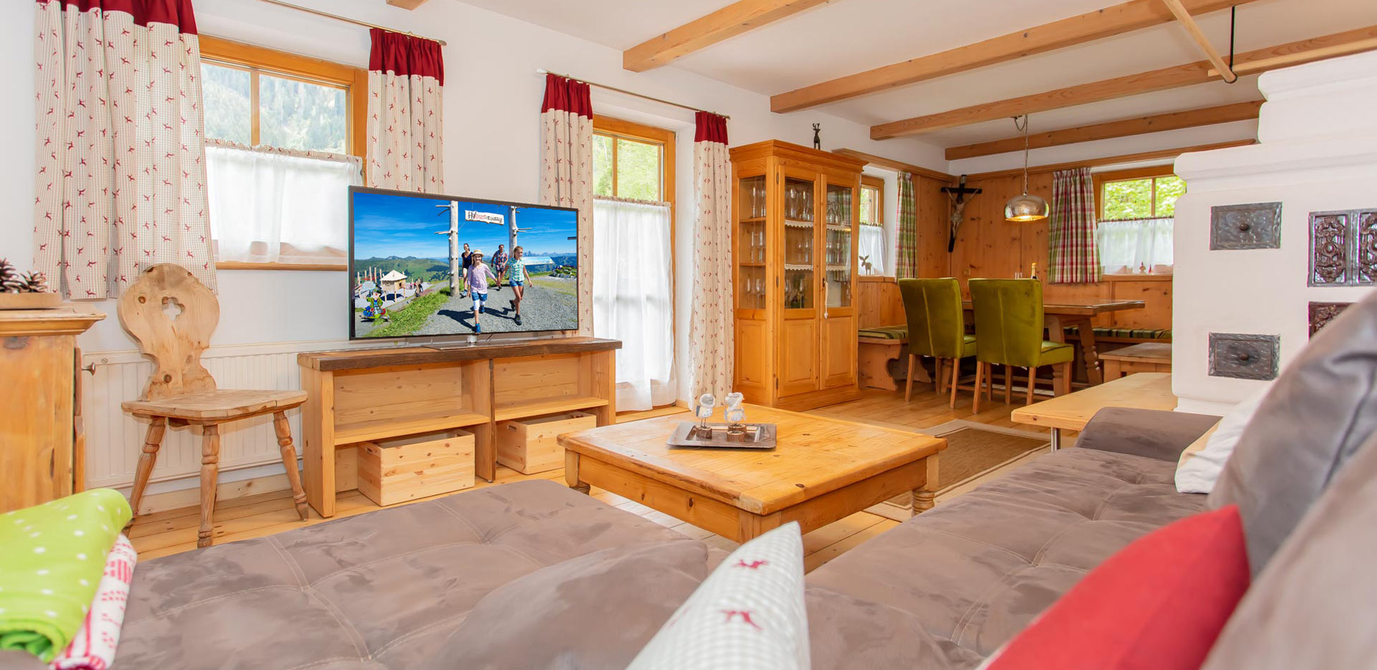Gemütlicher Wohnbereich eines Chalets in Saalbach‑Hinterglemm mit Holzmöbeln, Sofabereich, großem Flachbildfernseher auf Holzkonsole, traditionellem Kachelofen und Essbereich mit Holzstühlen und grünen Polstern.