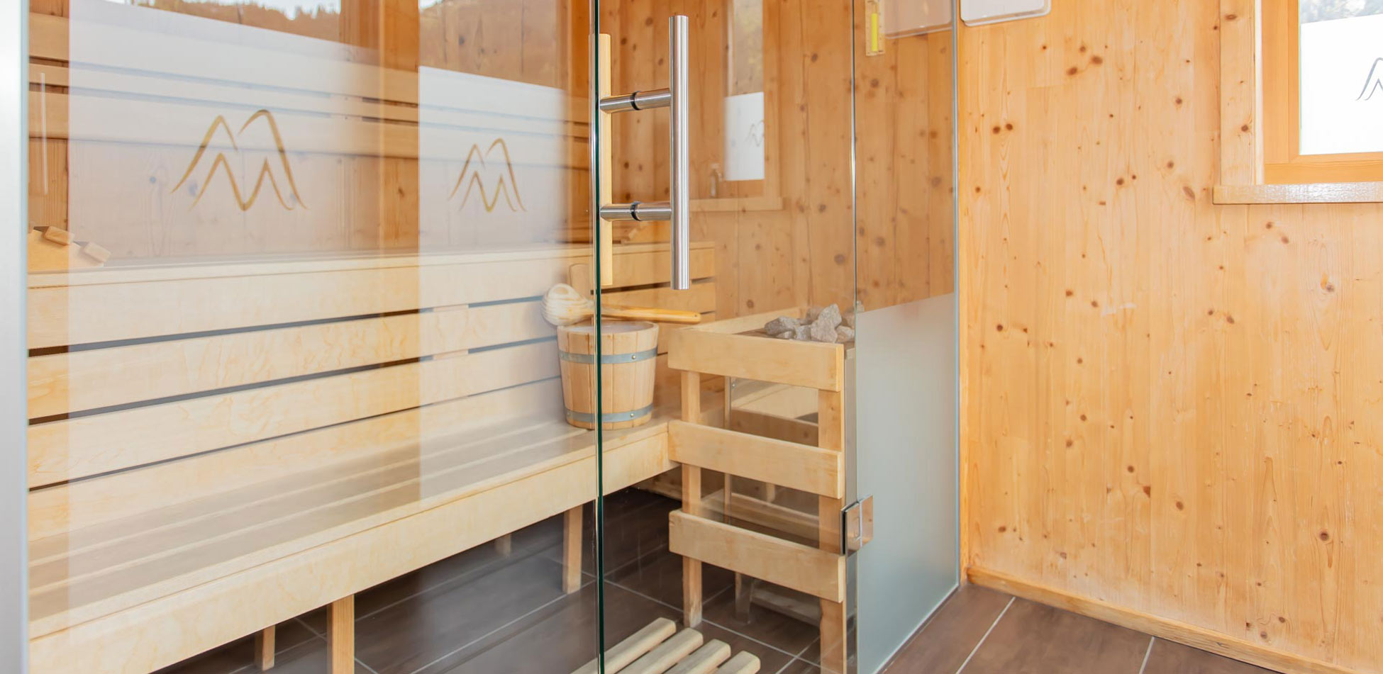 Saunabereich mit Holzbänken, Saunaofen mit Steinen und Glastür in einem Raum mit Holzverkleidung