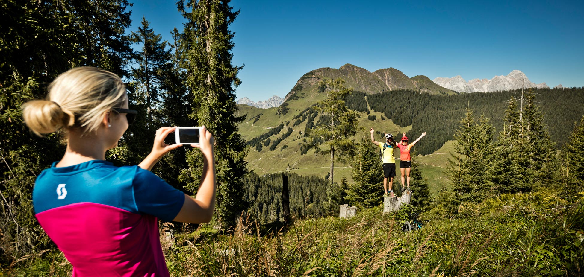 Frau mit blondem Dutt in blau-pinkem Sportshirt fotografiert zwei Personen mit Fahrradhelmen, die auf Baumstümpfen vor Berglandschaft posieren