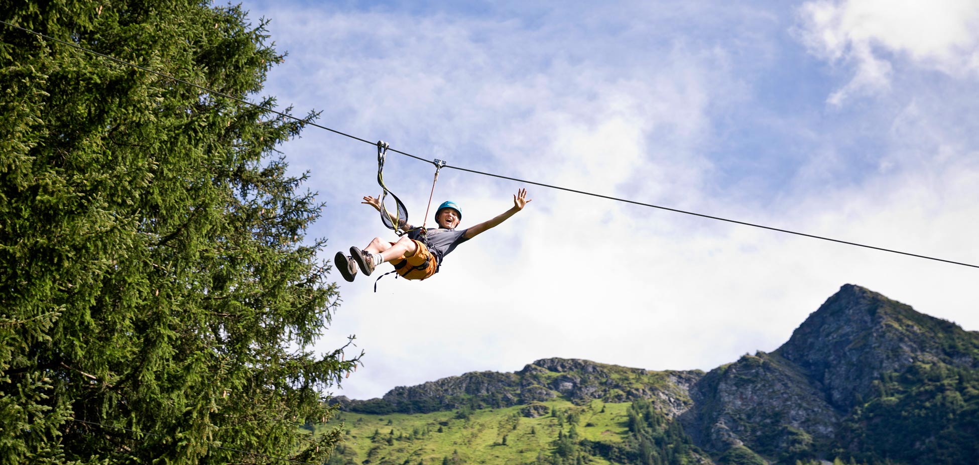 Person mit Helm und Klettergurt schwebt an einer Zipline über bewaldete Berge und grünes Tal