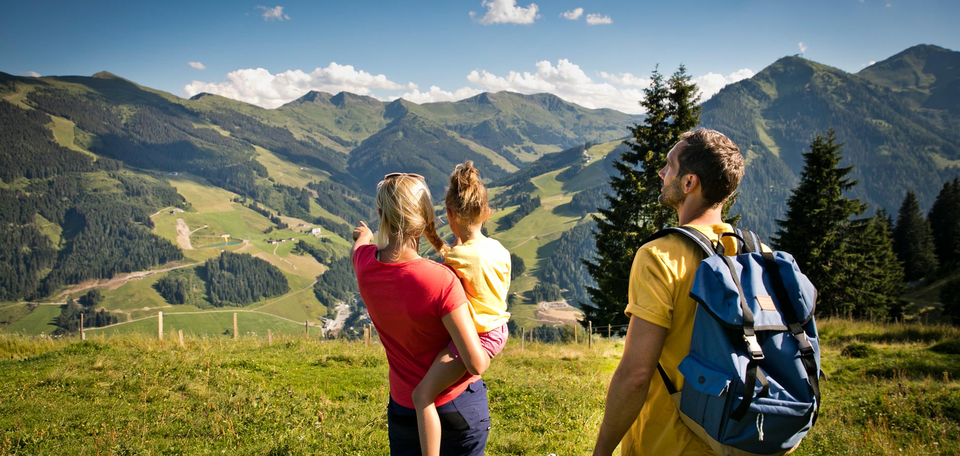 Drei Personen, eine Frau hält ein Kind, ein Mann mit Rucksack steht auf einer Wiese und blickt auf bewaldete Berge und Täler