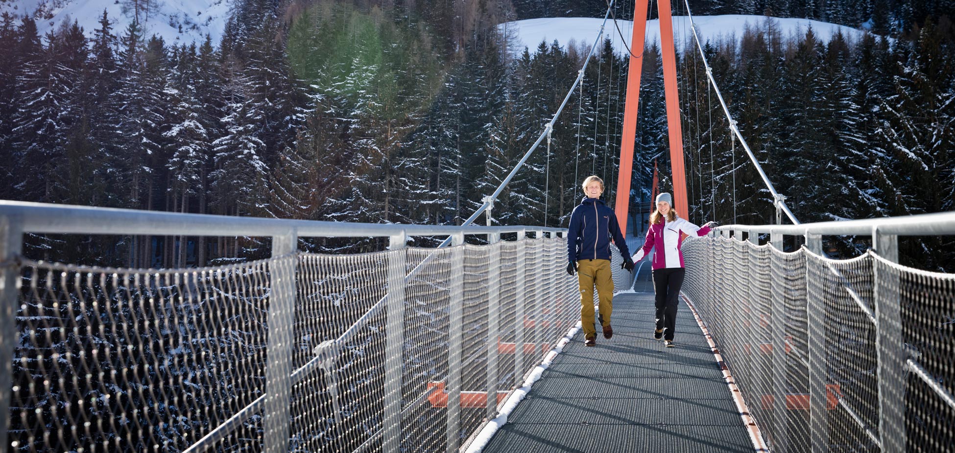 Zwei Personen gehen auf einer Hängebrücke in einem verschneiten Waldgebiet