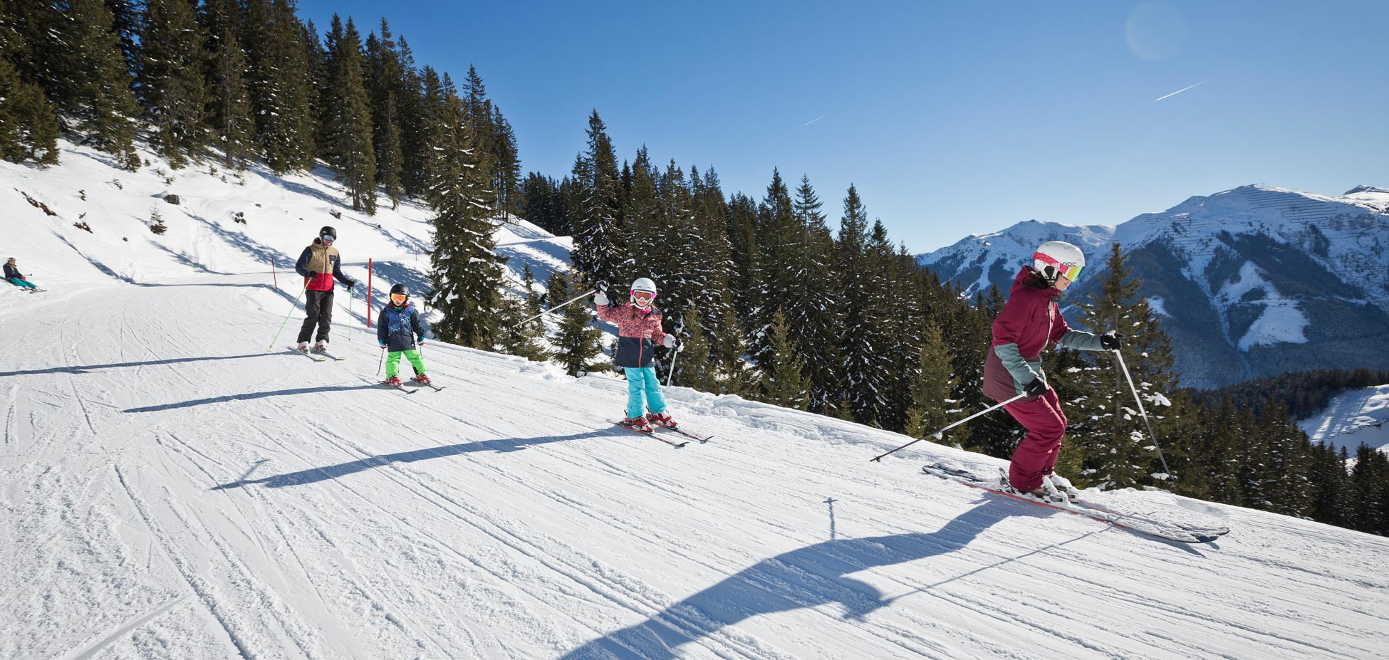 Gruppe von Skifahrern unterschiedlichen Alters auf einer schneebedeckten Piste vor bewaldeten Bergen und blauem Himmel