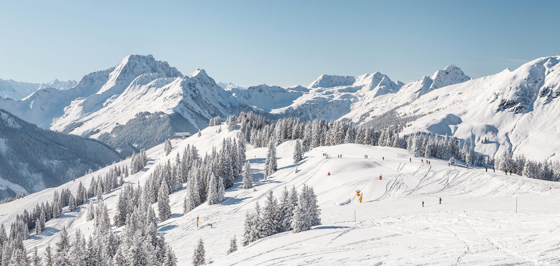 Verschneite Berglandschaft mit Tannen, Skifahrern auf Pisten und im Hintergrund schneebedeckte Gipfel