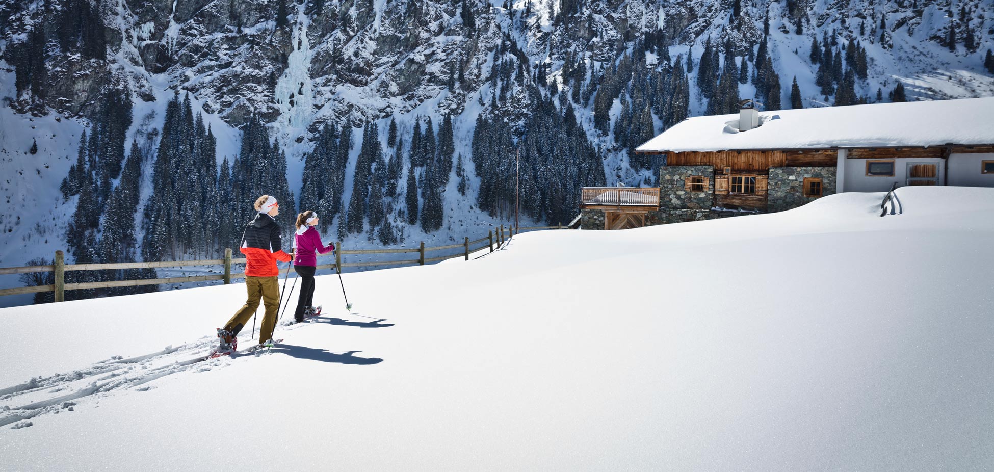 Zwei Personen mit Schneeschuhen gehen auf einer verschneiten Fläche auf eine Berghütte zu, im Hintergrund steile Felswände mit Nadelbäumen
