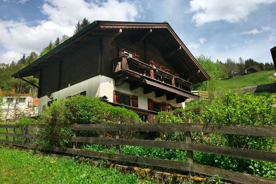 Two-story chalet with wooden upper floor and balcony surrounded by green bushes and wooden fence under a partly cloudy sky
