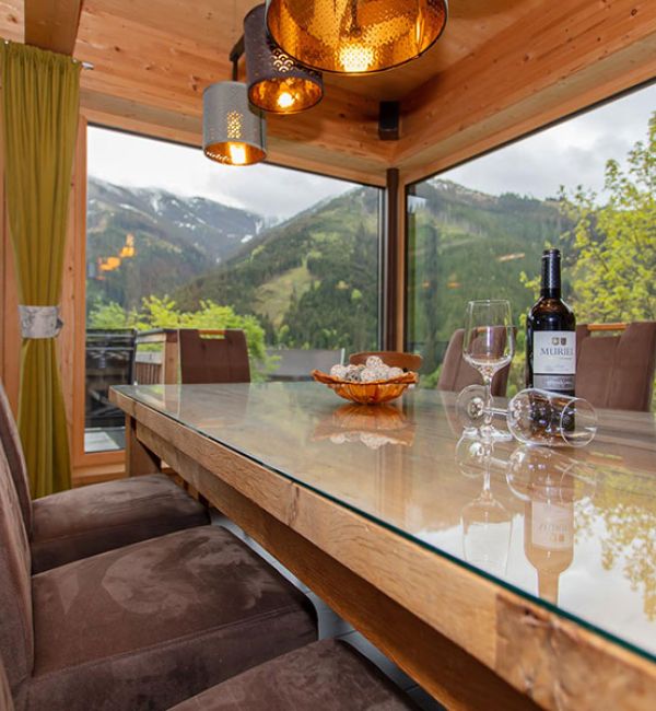 Essbereich in einem Chalet mit langem Holztisch und bequemen Stühlen vor großen Panoramafenstern mit Blick auf grüne Berge und Almwiesen in der Salzburger Bergwelt bei Viehhofen/Saalbach‑Hinterglemm.