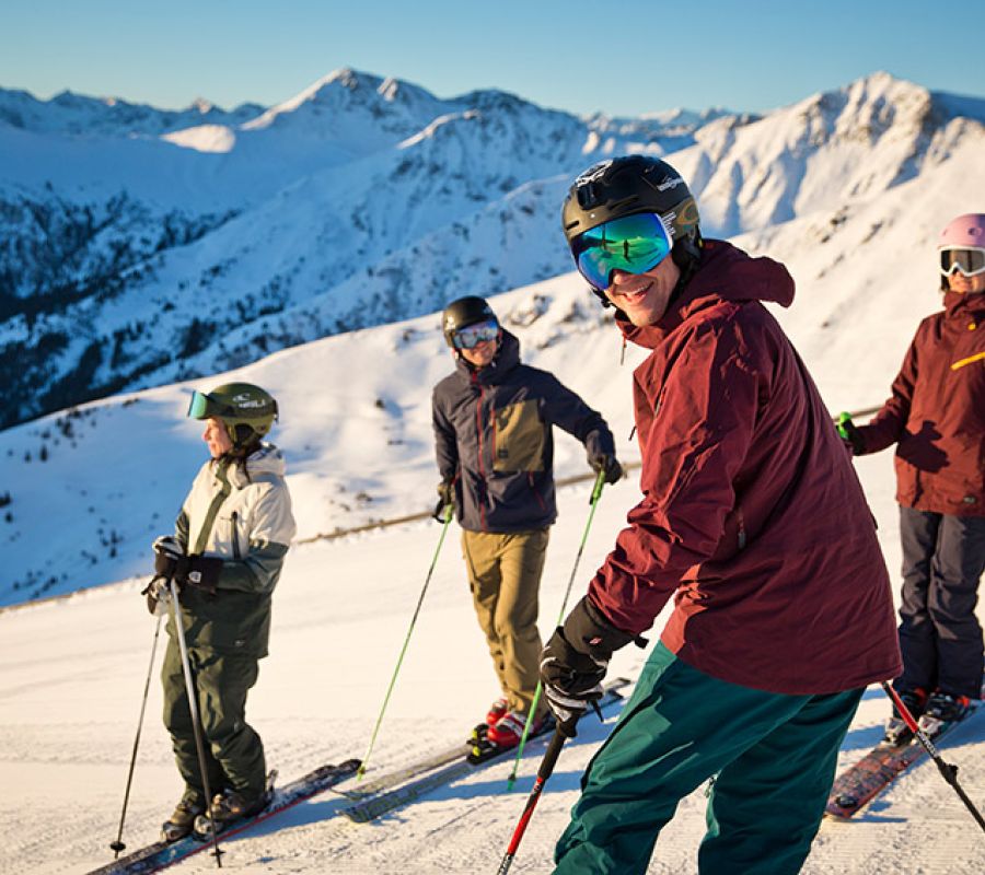 Vier Skifahrer stehen auf einer schneebedeckten Piste vor einer Bergkulisse