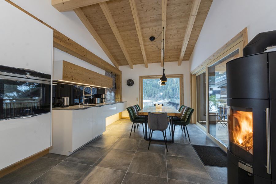 Moderne Küche mit weißer Einbauküche, Esstisch mit Stühlen vor großem Fenster und brennendem Kaminofen in Holzdecke mit sichtbaren Balken