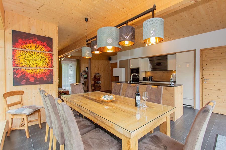 Essbereich mit großem Holztisch, mehreren gepolsterten Stühlen, moderner Küchenzeile im Hintergrund und vier hängenden Lampenschirmen