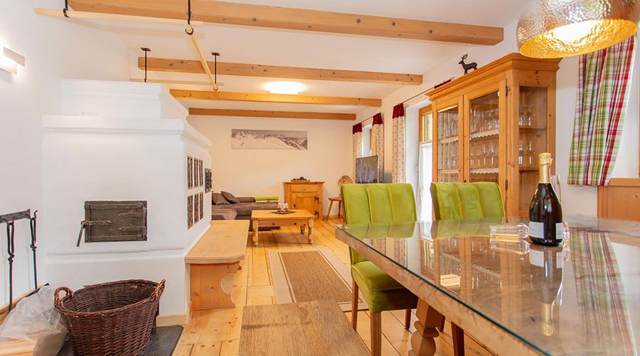 Wohnzimmer mit Holzboden, weißem Kachelofen, grün gepolsterten Stühlen, Glastisch und Holzvitrine mit Gläsern