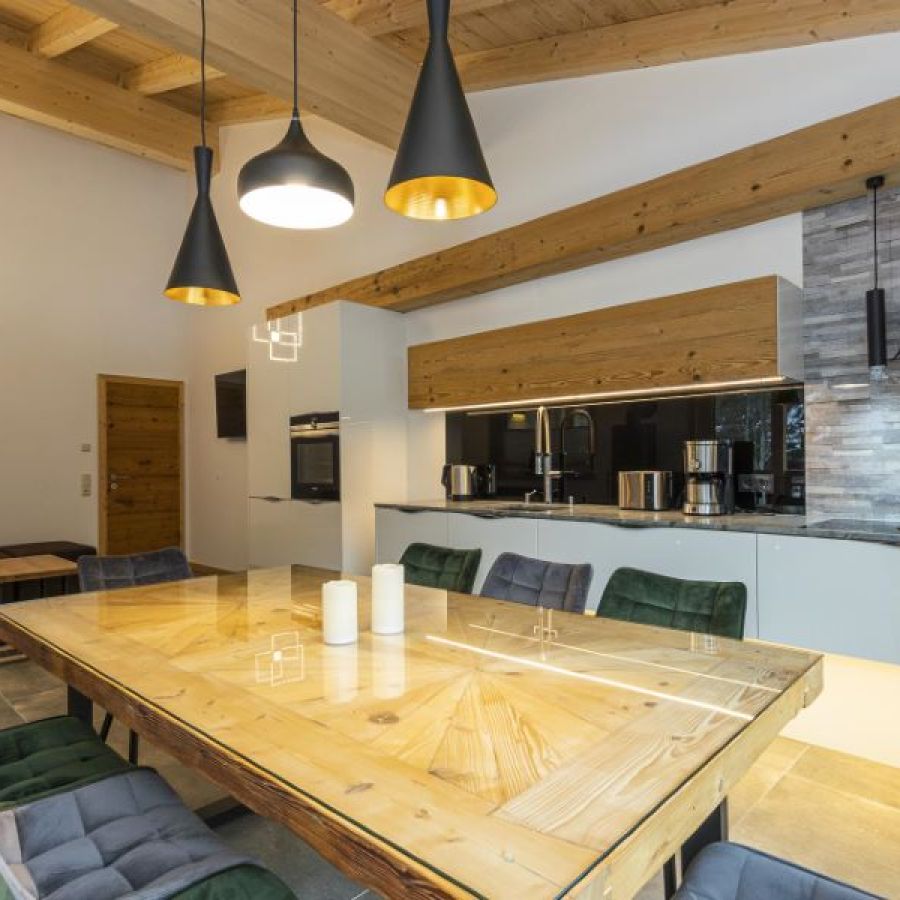 Moderner Essbereich mit großem Holztisch, gepolsterten Stühlen, offener Küche mit Holzfronten und drei hängenden Lampen unter sichtbaren Holzbalken