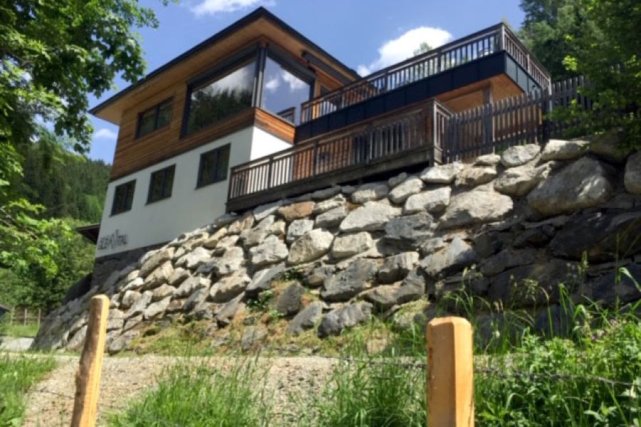 House with large windows and wooden balcony on a stone retaining wall surrounded by trees and grass under a partly cloudy sky