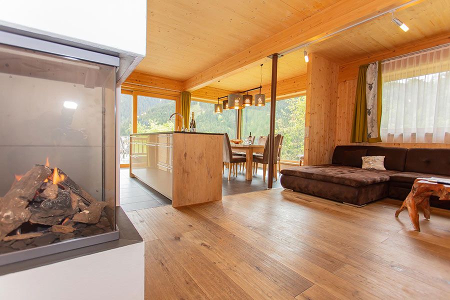 Wohnzimmer mit Holzboden, brauner Eckcouch, Esstisch mit Stühlen vor großen Fenstern und moderner Küche mit Kochinsel sowie Kamin im Vordergrund