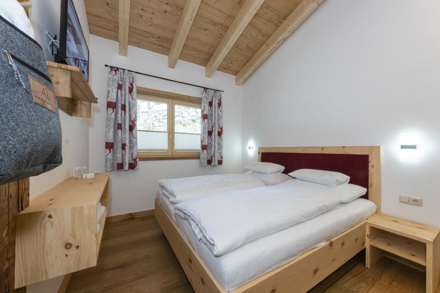 Schlafzimmer mit Holzbett, weißer Bettwäsche, Holzboden, Holzwandregal und Fenster mit gemusterten Vorhängen unter einer sichtbaren Holzbalkendecke