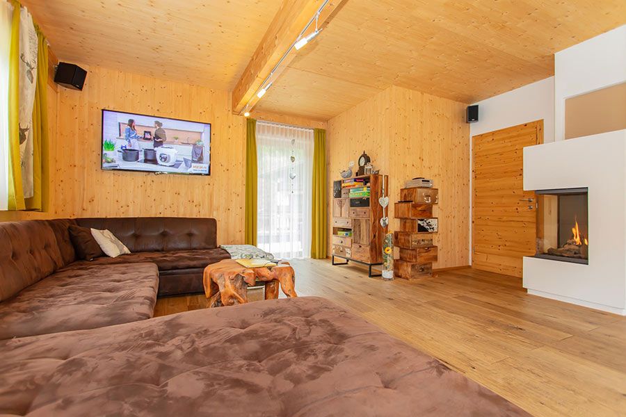Wohnzimmer mit brauner Eckcouch, Holzverkleidung an Wänden und Decke, Kamin, Fernseher an der Wand und Holzkommode mit Dekoration