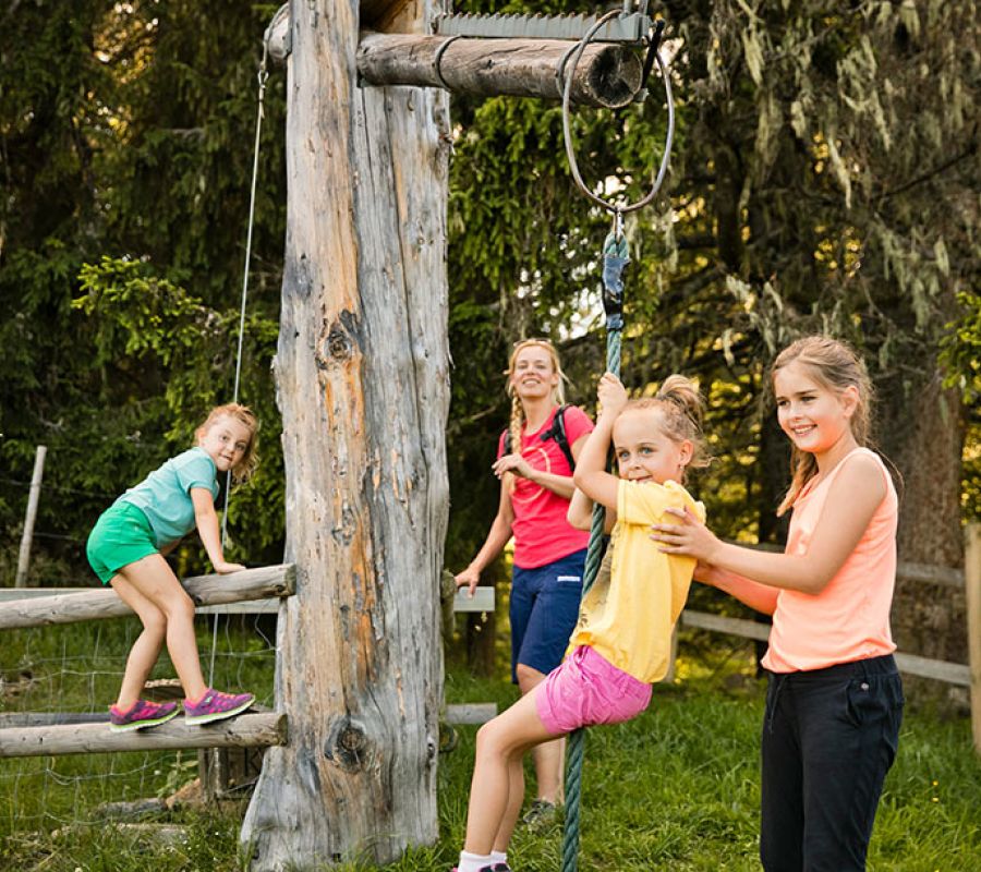Vier Kinder spielen an einem Klettergerüst aus Holz im Freien, eines klettert auf einem Zaun, ein anderes hält sich an einem Seil fest, zwei weitere stehen daneben