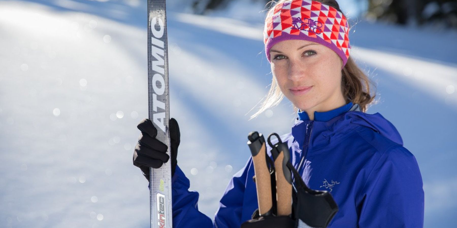 Person in blue jacket and patterned headband holding cross-country skis and poles in snowy landscape