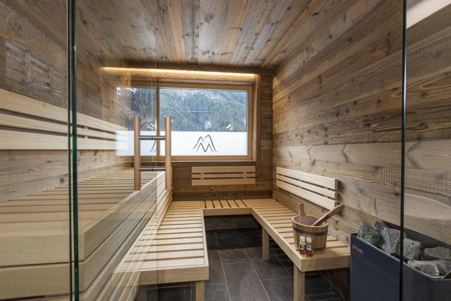 Saunaraum mit Holzwänden und -bänken neben einem Waschbereich mit zwei weißen Waschbecken auf einer Holzablage, Glastür