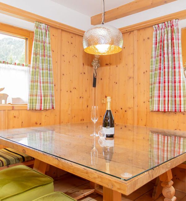 Wood-paneled dining nook with a glass-topped table, green cushioned chairs, plaid curtains, and a hanging lamp