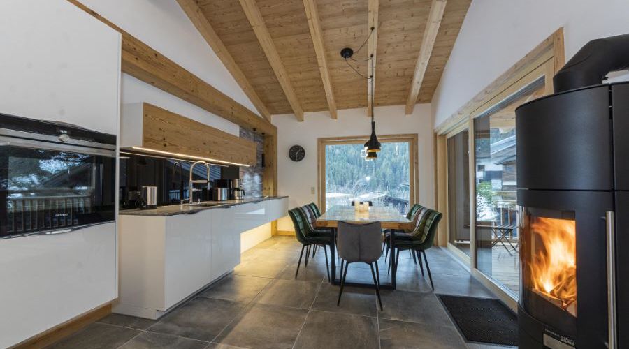 Offene Küche mit weißer Küchenzeile, Esstisch mit Stühlen vor großem Fenster und brennendem Kaminofen in modernem Holzhaus