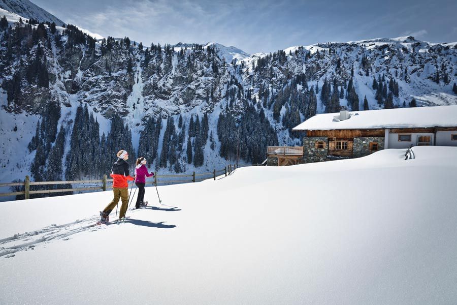 Zwei Personen beim Skitourengehen auf verschneitem Hang vor bewaldetem Bergpanorama und Hütte
