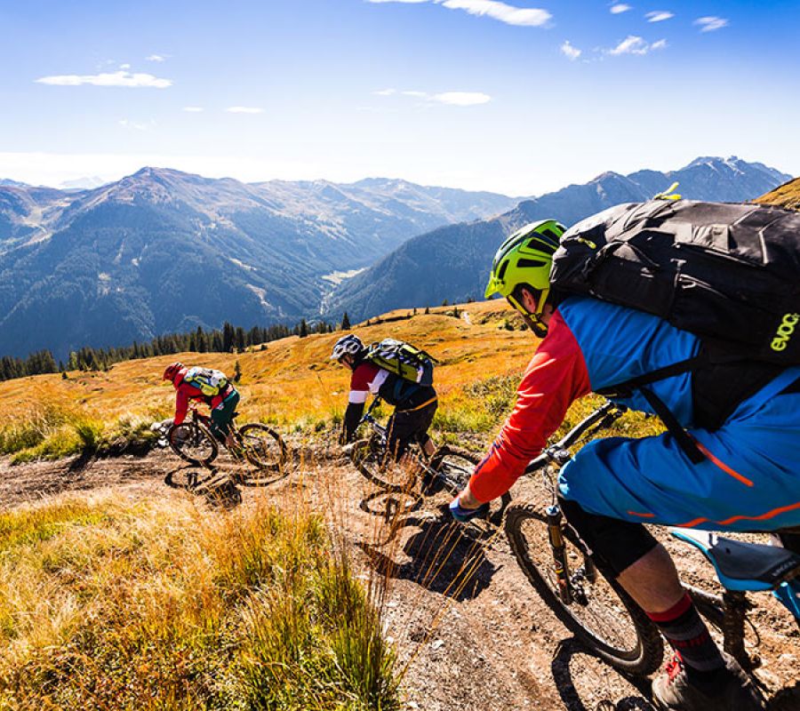 Drei Mountainbiker mit Helmen und Rucksäcken fahren auf einem schmalen Pfad durch eine bergige Landschaft mit herbstlicher Vegetation und Bergen im Hintergrund