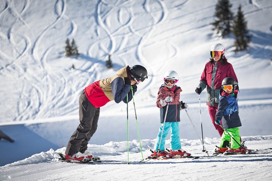 Vier Personen in Skibekleidung mit Helmen und Skiern stehen auf einer Schneepiste vor verschneiten Hängen mit Skispuren