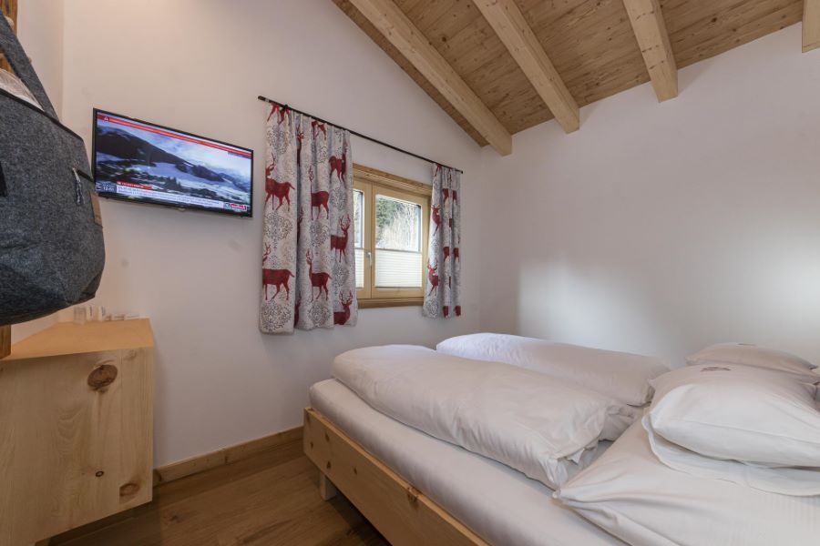 Kleines Schlafzimmer mit Holzbett, weißen Bettdecken, Fenster mit Gardinen mit Hirschmotiv und Wandfernseher unter schräger Holzbalkendecke