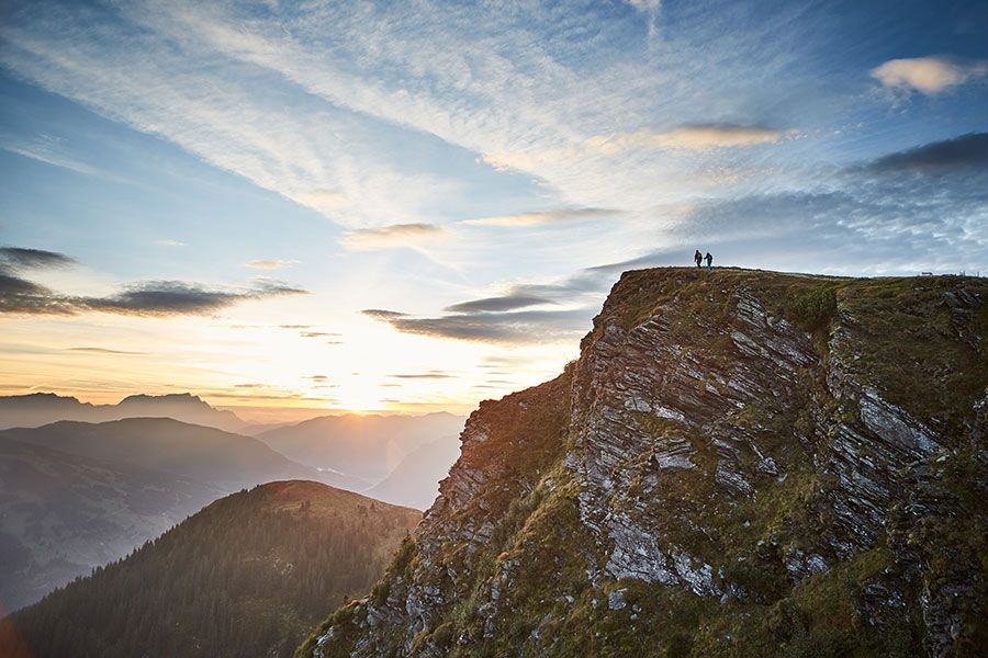Zwei Personen stehen auf einem felsigen Berggipfel bei Sonnenuntergang mit Berglandschaft im Hintergrund