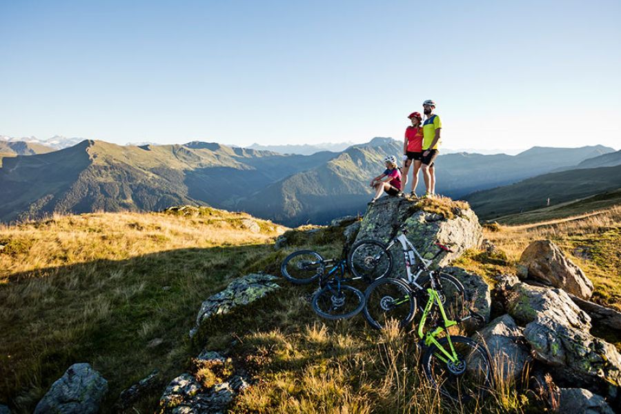 Drei Radfahrer mit Helmen stehen und sitzen auf einem Felsen in einer Berglandschaft mit mehreren Mountainbikes im Vordergrund