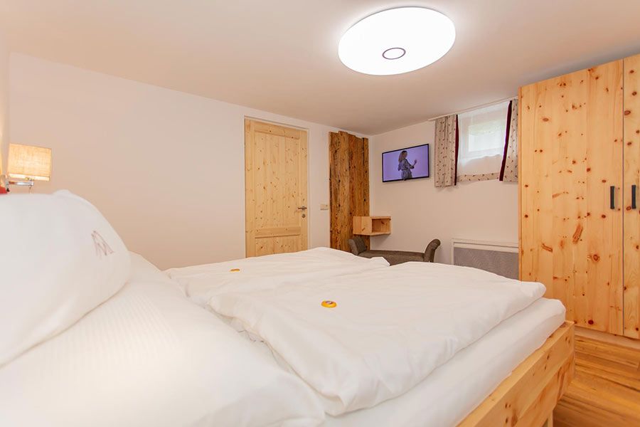 Bedroom with wooden bed, wardrobe, door, small window with curtains, wall-mounted TV, and ceiling light