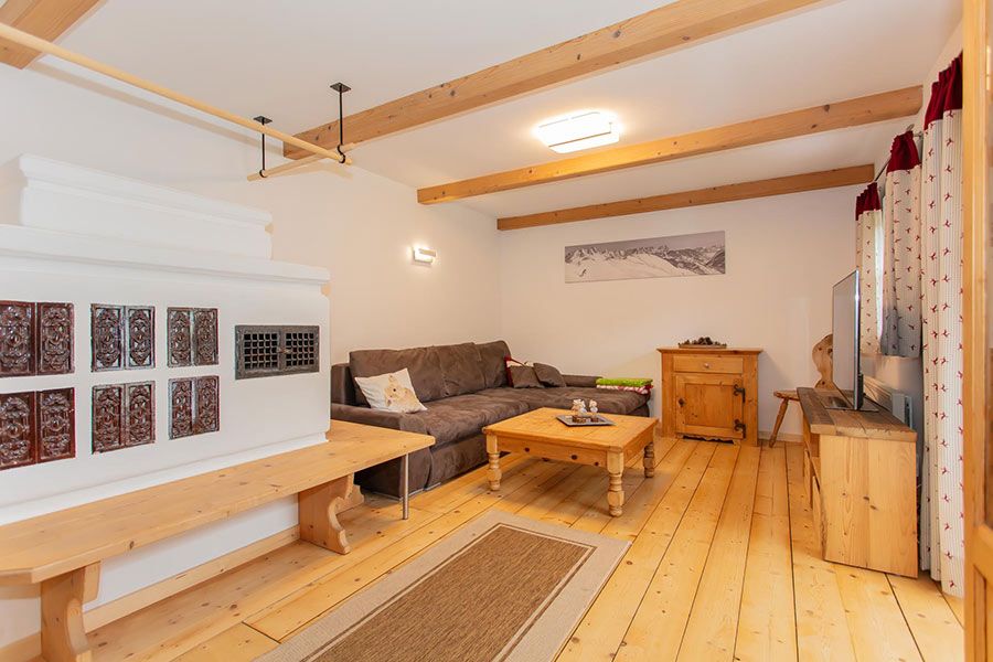 Living room with wooden floor and ceiling beams, brown sofa, wooden coffee table, TV on wooden stand, and white tiled stove with wooden bench
