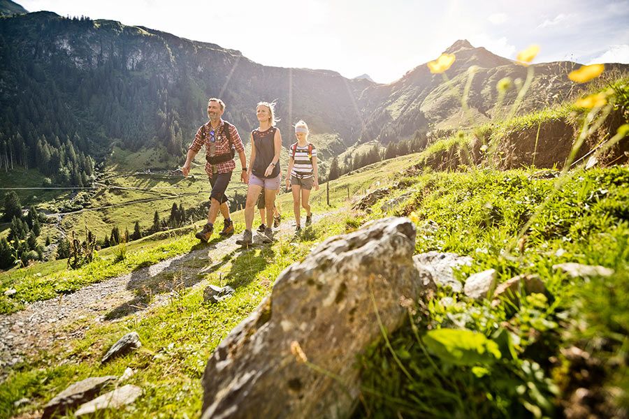 Drei Wanderer auf einem Bergpfad mit grüner Wiese und Bergen im Hintergrund