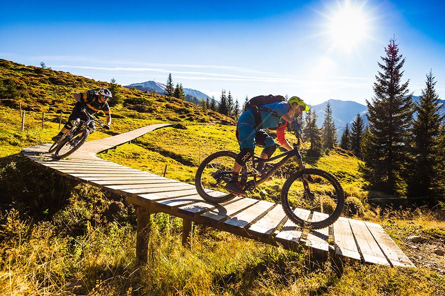 Zwei Mountainbiker fahren auf einem schmalen Holzsteg in bergiger Landschaft bei Sonnenschein