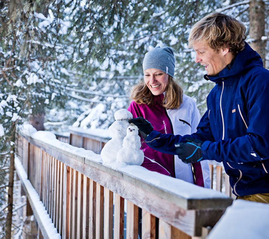 Zwei Personen bauen auf einem schneebedeckten Holzgeländer kleine Schneemänner