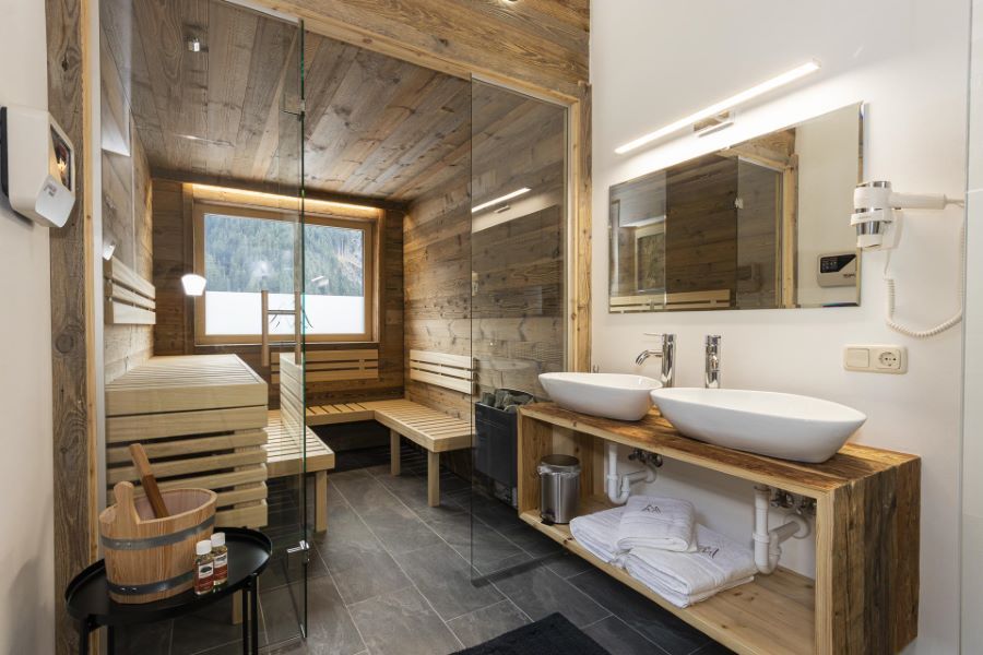 Saunaraum mit Holzwänden und -bänken neben einem Waschbereich mit zwei weißen Waschbecken auf einer Holzablage und großem Spiegel