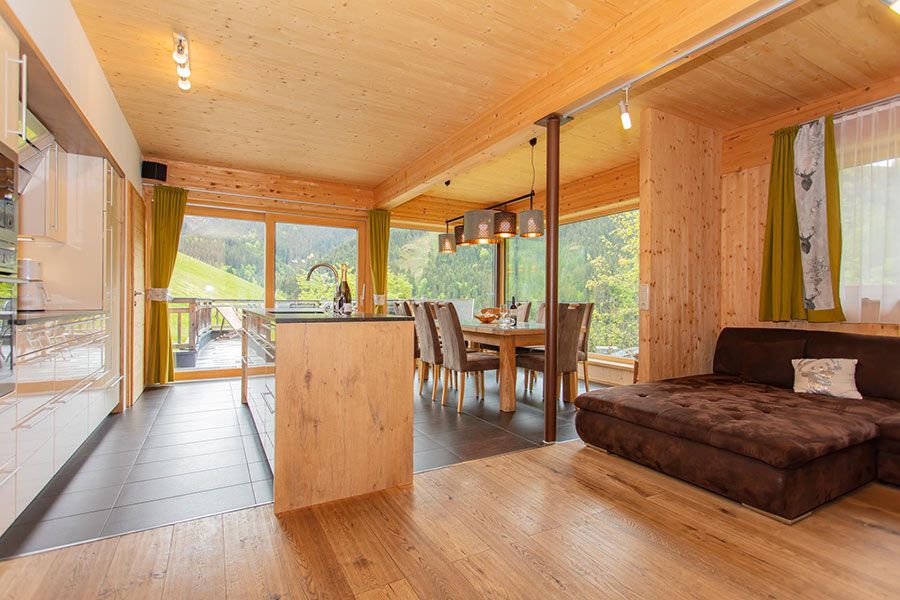 Open-plan wooden interior with kitchen island, dining table with chairs, and brown upholstered sofa near window with green curtains