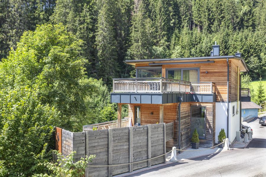 Modernes Holz‑Chalet an einem sonnigen Sommertag am Waldrand in Viehhofen bei Saalbach‑Hinterglemm mit großer Terrasse und umliegenden grünen Bäumen und Bergen im Hintergrund.