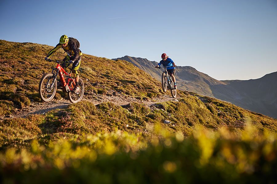 Zwei Mountainbiker fahren auf einem schmalen Bergpfad mit grasbewachsenen Hängen und Bergen im Hintergrund