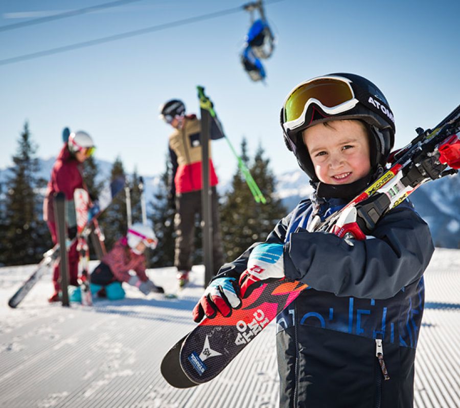 Kind mit Skihelm und Skibrille trägt Skier auf der Schulter vor verschneiter Berglandschaft und weiteren Skifahrern