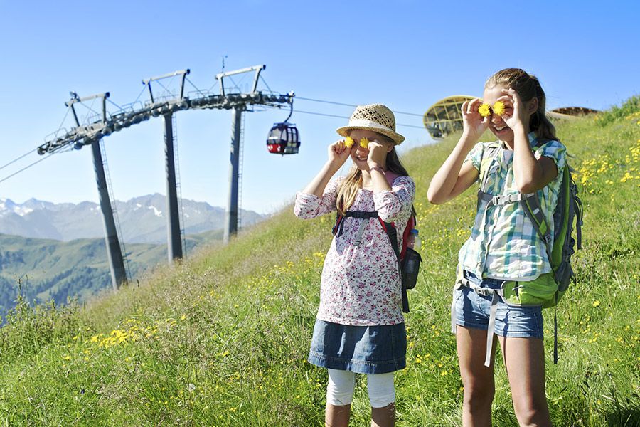 Zwei Mädchen mit Rucksäcken und Sonnenhüten stehen auf einer Blumenwiese vor einer Seilbahn in den Bergen
