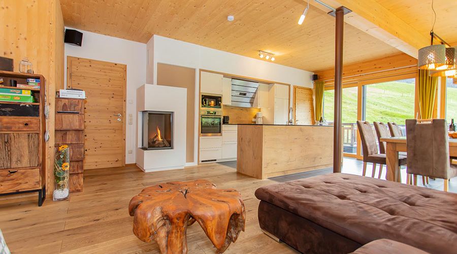 Offener Wohnraum mit Holzboden, Kamin, moderner Küche, rustikalem Baumstamm-Tisch und großem Esstisch vor bodentiefen Fenstern mit Blick ins Grüne