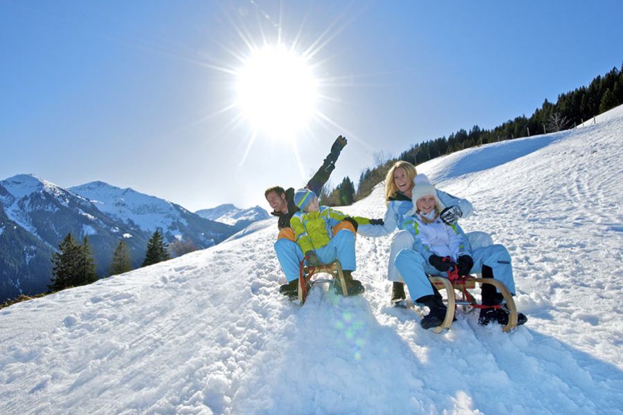 Familie mit zwei Kindern sitzt auf zwei Schlitten auf schneebedecktem Hang vor Bergkulisse bei Sonnenschein