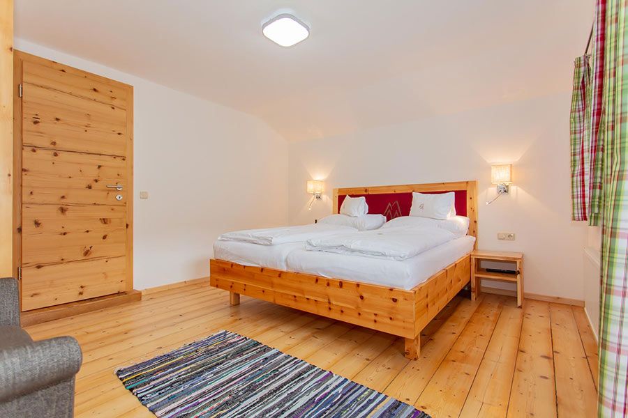 Bedroom with wooden floor and door, double bed with white bedding and red headboard, two wall lamps, small wooden nightstand, striped rug, and plaid curtains