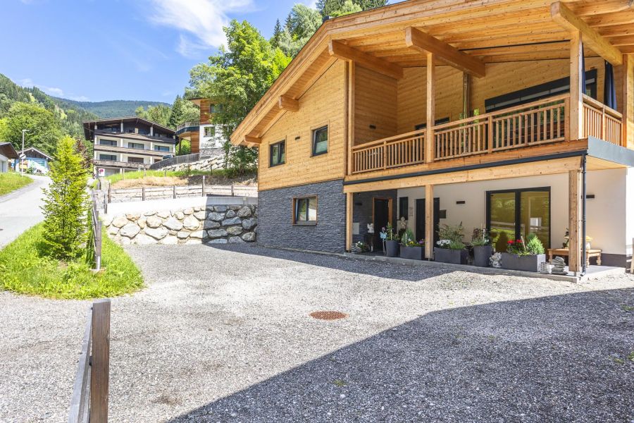 Modernes Chalet in den Alpen der Ferienregion Saalbach‑Hinterglemm/Viehhofen mit großer Holzterrasse, Panoramafenstern und Blick auf grüne Wiesen und Berge an einem sonnigen Tag.