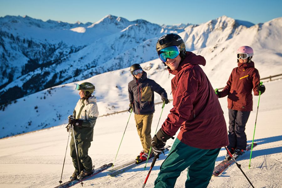 Vier Skifahrer mit Helmen und Skistöcken stehen auf schneebedeckter Piste vor Bergpanorama